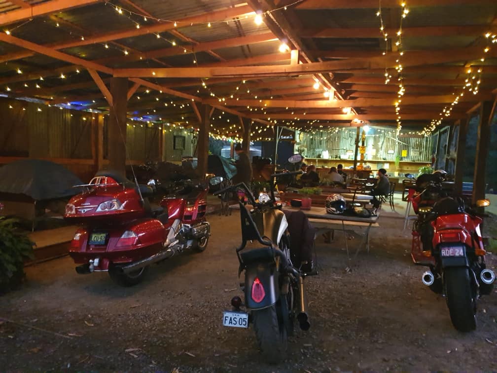 Several motorcycles are parked under a rustic, open-air timber structure illuminated by warm string lights