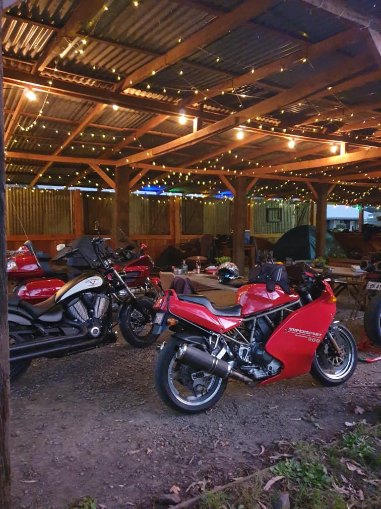A red Ducati Supersport 900 and a black cruiser motorcycle are parked under a rustic timber shelter decorated with warm string lights at night.