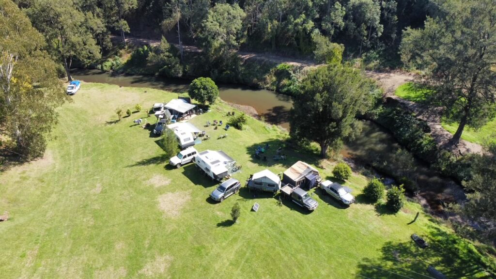 A line of camper trailers, SUVs, and trucks parked on a grassy field beside a winding river, surrounded by lush green trees.