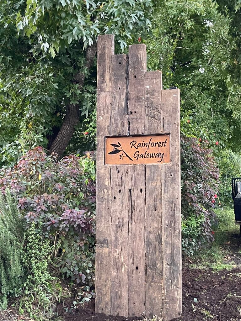 A rustic wooden sign made of vertical timber planks displays a metal plaque that reads "Rainforest Gateway" with a leaf illustration