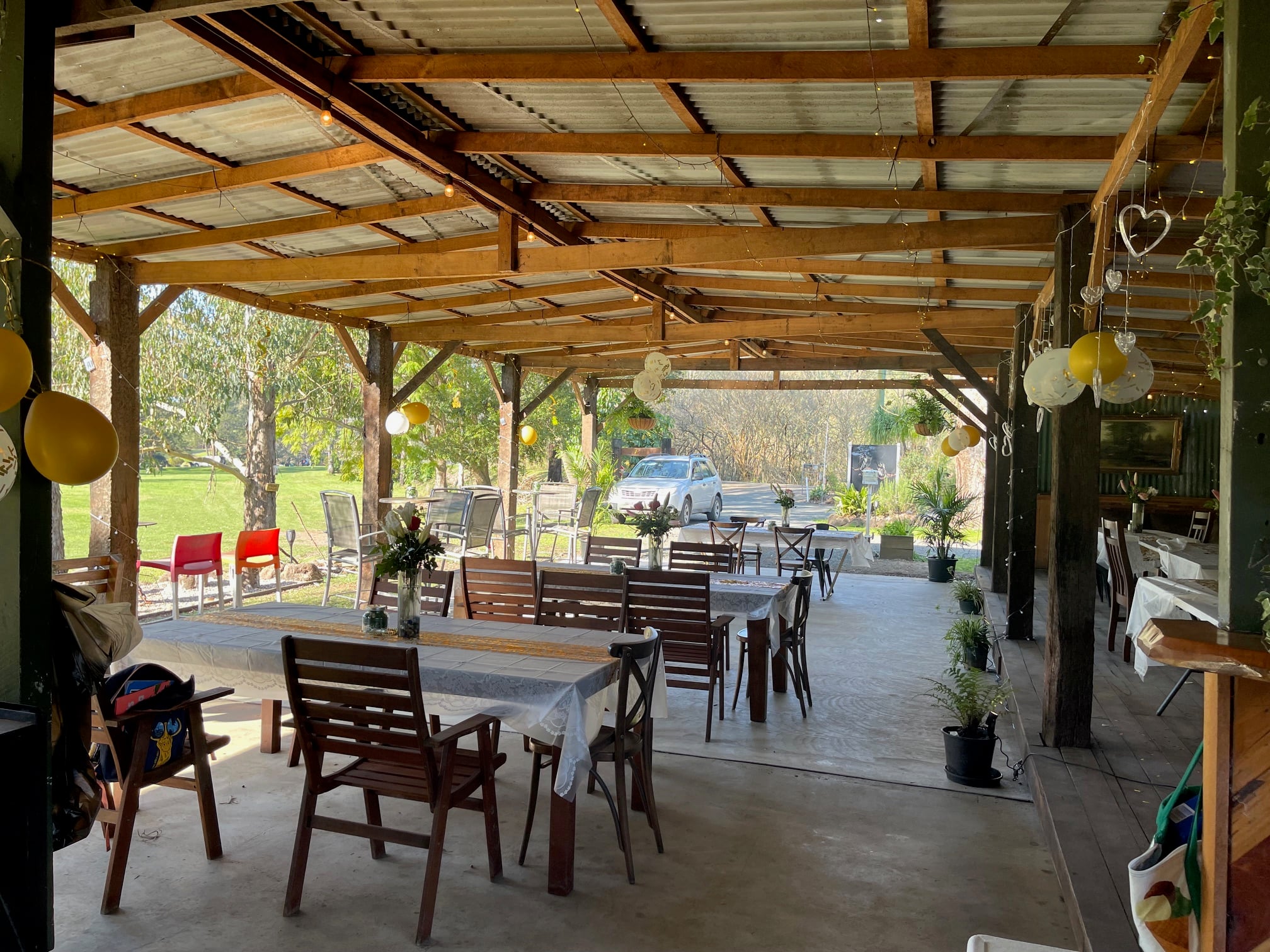A long, shaded outdoor event space decorated with yellow balloons and string lights, featuring several wooden dining tables set with white linens.