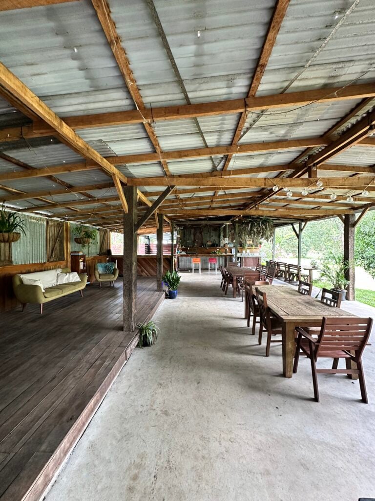 A wide, rustic outdoor pavilion featuring a corrugated metal roof and timber beams, furnished with wooden dining tables, a green sofa on a raised deck, and a bar area in the background.