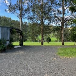 A gravel courtyard sits beside a corrugated metal shed, opening onto a bright green field framed by tall eucalyptus trees.
