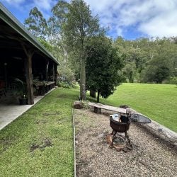 A rustic outdoor area featuring a metal fire pit on a gravel patch next to a long wooden bench, overlooking a large green lawn and a forested hillside.