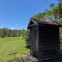 A rustic, weathered wooden outhouse with a corrugated metal roof stands on a small timber platform in a lush green field under a clear blue sky.