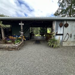 The exterior view of the open-fronted shed showing a gravel path leading to the kitchen and storage area, with kayaks stored on the left and vintage tools mounted on the corrugated metal exterior.