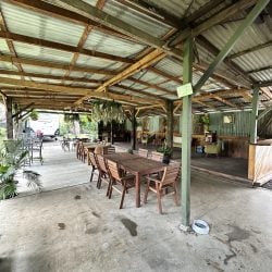 An interior view of a spacious, rustic pavilion featuring wooden dining tables and chairs on a concrete floor, with a raised wooden lounge area in the background.