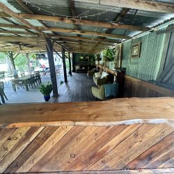 A rustic, indoor-outdoor lounge area viewed from behind a live-edge wooden bar, featuring corrugated metal walls.