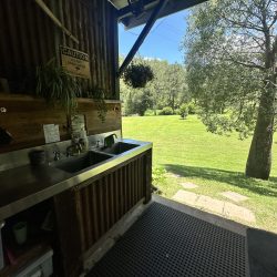 A stainless steel double sink is built into a corrugated metal and wood vanity under a sheltered area.