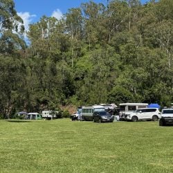 Several campers, SUVs, and a tent are parked in a wide, green meadow at the base of a densely forested hill.