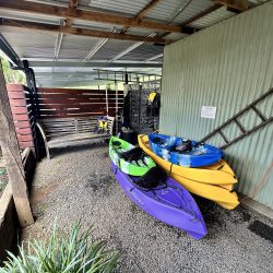 A gravel-covered storage area under a corrugated metal roof containing several colourful kayaks