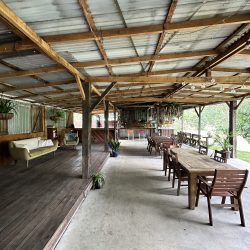 A wide, rustic outdoor pavilion featuring a corrugated metal roof and timber beams, furnished with wooden dining tables, a green sofa on a raised deck, and a bar area in the background.