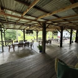 A view from a wooden deck looking into a rustic, open-air pavilion with several wooden dining sets under a corrugated metal roof